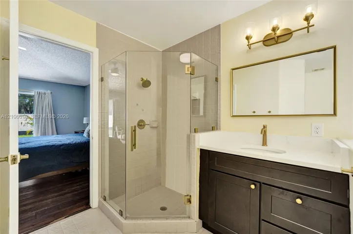 Bathroom featuring a corner glass-enclosed shower, dark wood-finish vanity with a light stone-style countertop, rectangular mirror with gold-tone frame, and matching gold-tone plumbing fixtures