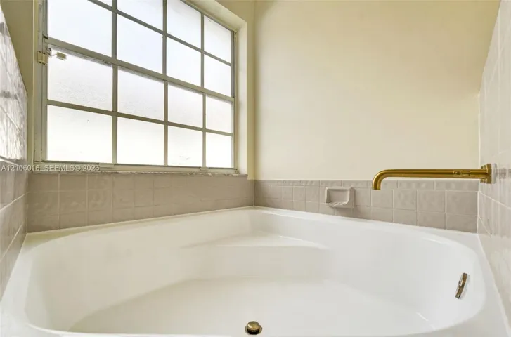 Corner soaking tub featuring a metallic wall-mounted faucet, ceramic tile surround, and a multi-pane grid window