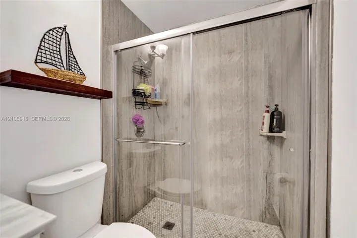 Walk-in shower featuring large-format stone-look wall tile, hexagonal mosaic floor tile, and a sliding glass enclosure with a metallic frame, accompanied by a mounted wood-finish display shelf