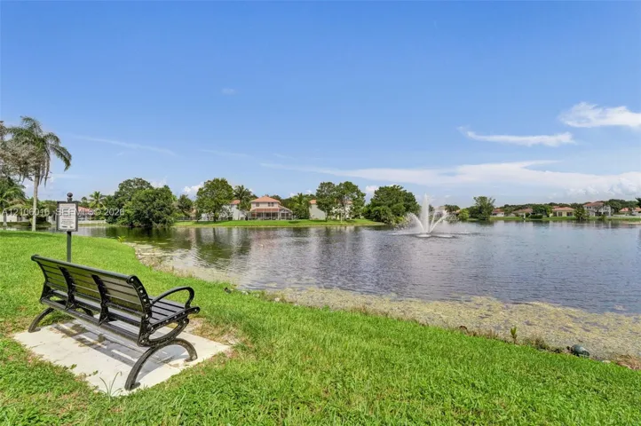 Waterfront setting featuring a concrete pad, metal bench, mature landscaping, and a central fountain