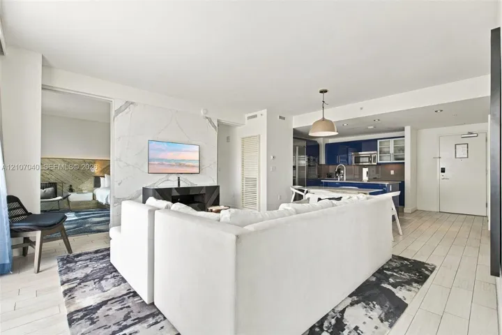 Open-concept living space featuring large-format tile flooring, a floor-to-ceiling stone-surround wall, glossy navy cabinetry, integrated kitchen appliances, and a polished metal pendant light fixture
