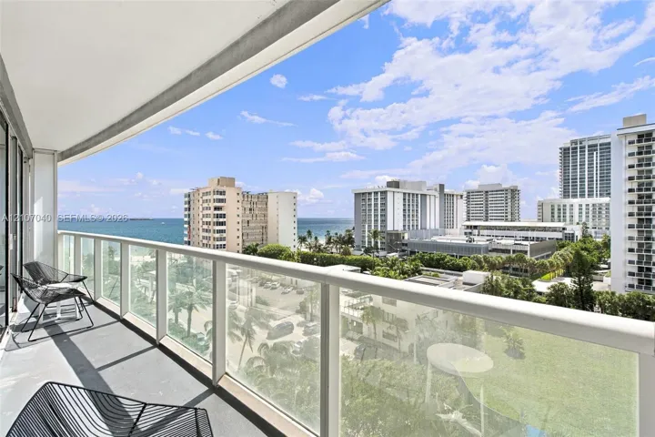 Private covered balcony featuring a glass panel railing, concrete flooring, and panoramic coastal and urban scenery