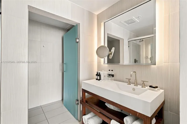 Open-concept vanity featuring a white composite countertop, integrated rectangular sink, metal widespread faucet, wood-finish shelving, and wall-mounted mirror with integrated lighting, finished with floor-to-ceiling tile work and a frosted glass door