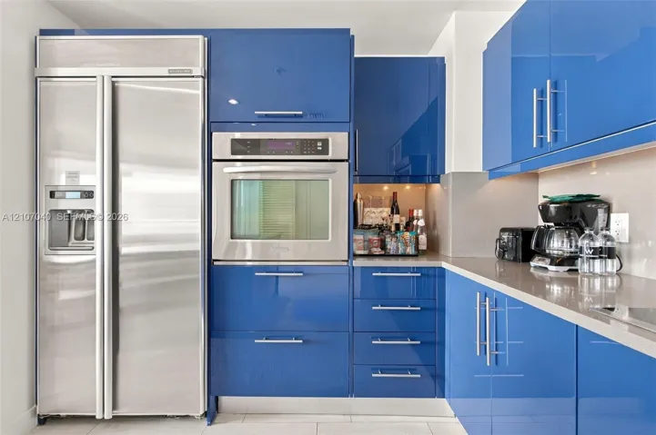 Kitchen featuring high-gloss blue cabinetry with polished metal hardware, integrated stainless steel side-by-side refrigerator, built-in wall oven, and neutral solid-surface countertops