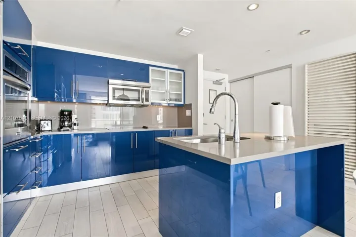 Glossy blue cabinetry, solid surface countertops, center island with an integrated sink, stainless steel microwave, and wood-finish tile flooring
