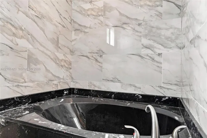 Built-in soaking tub with a dark stone-finish surround, complemented by floor-to-ceiling wall tile featuring a veined pattern and a metallic faucet fixture
