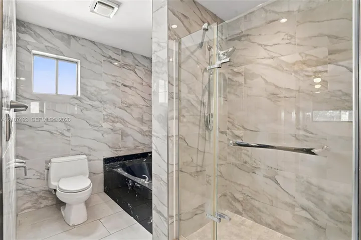 Bathroom featuring floor-to-ceiling veined tile, a glass-enclosed shower, a black stone-finish bathtub surround, and a white porcelain toilet