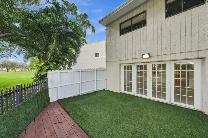 Exterior featuring vertical siding, four-panel glass doors, synthetic turf lawn, wood-finish decking, and partial privacy fencing