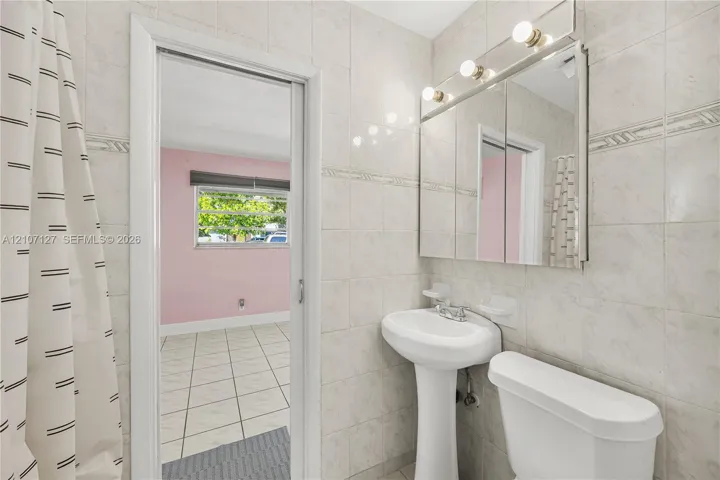Full-height tile wall surround, white porcelain pedestal sink, mirrored medicine cabinet with integrated vanity lighting, and standard toilet