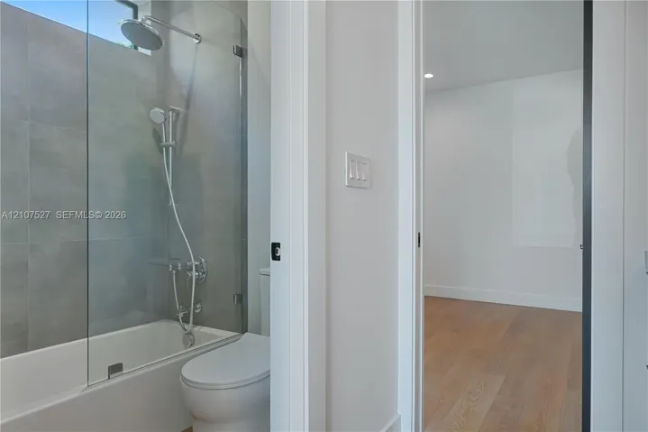 Bathroom featuring large-format grey tile surround, glass panel shower screen, rainfall shower head, and wood-finish flooring in the room beyond
