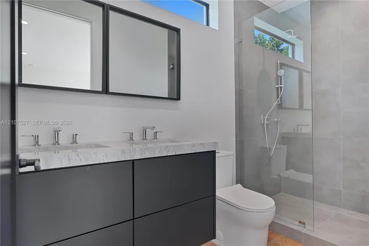 Floating dual-sink vanity with a stone countertop, framed mirrors, glass-enclosed shower with large-format tile, and rectangular transom windows