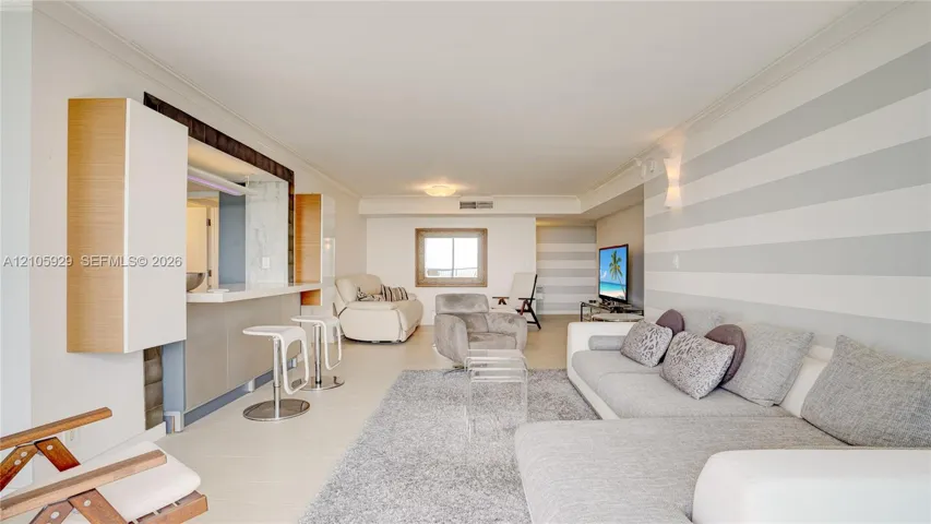 Living area featuring a service pass-through with a breakfast bar, neutral-tone striped wall treatments, crown molding, and large-format tile flooring
