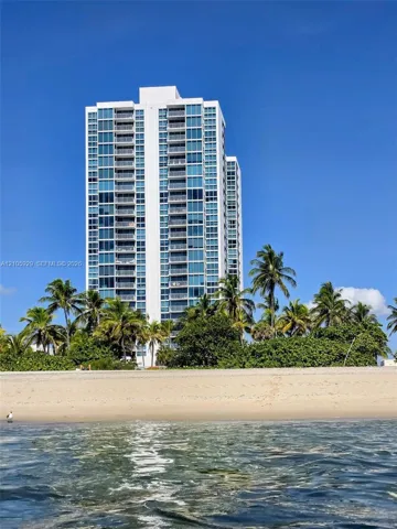 High-rise residential tower featuring a glass facade, private balconies, and direct beachfront access, surrounded by mature palm trees- view from the ocean