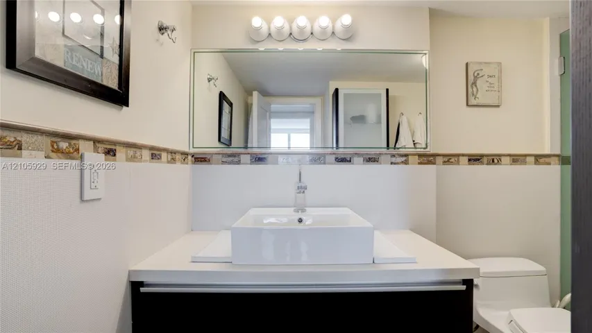Vanity featuring a rectangular vessel sink, solid-surface countertop, decorative tile backsplash, wall-mounted mirror, and multi-bulb light fixture