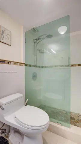 Walk-in shower featuring a frameless glass partition, wall-mounted rainfall showerhead, decorative tile border, and mosaic stone-finish floor