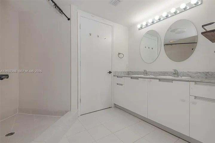 Bathroom featuring a dual-sink vanity with stone countertop, twin circular mirrors, a multi-bulb light fixture, and a walk-in shower with tile surround