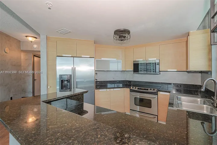 Kitchen featuring flat-panel light wood cabinetry, dark stone countertops, stainless steel appliances, breakfast bar, and stainless steel dual-basin sink