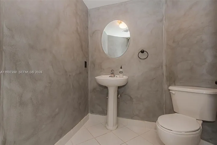 Textured concrete-finish walls, white ceramic pedestal sink, oval wall-mounted mirror, matte black towel ring, and ceramic tile flooring