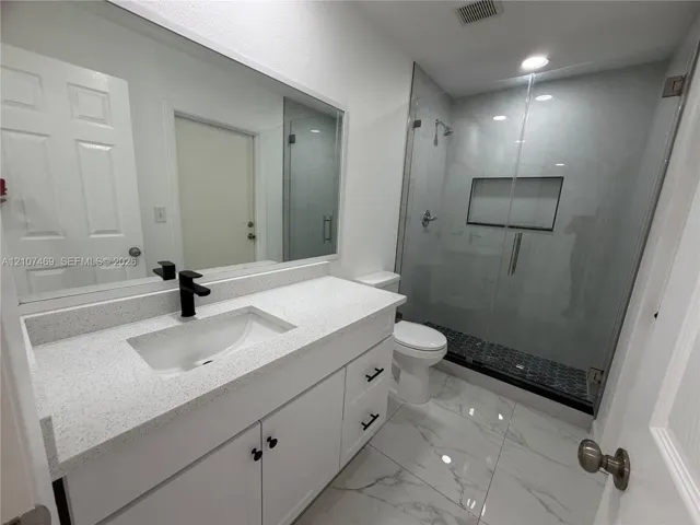 Bathroom featuring a white vanity with a light-toned solid surface countertop, matte black faucet, large wall-mounted mirror, glass-enclosed shower with a built-in recessed niche, and large-format tile flooring