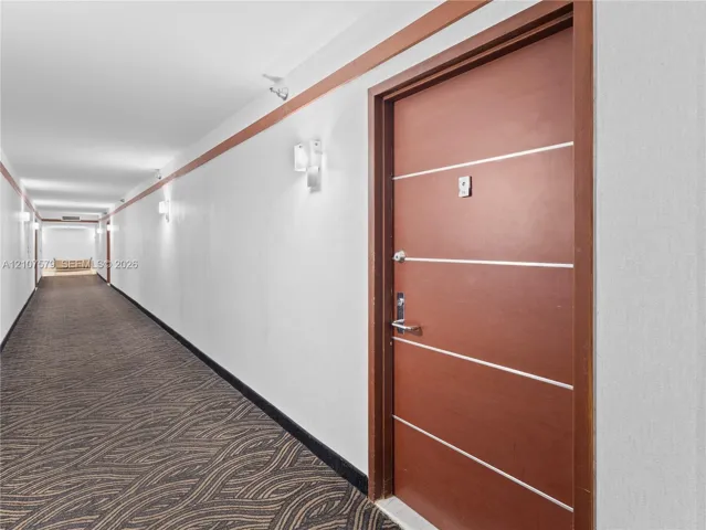 Interior hallway featuring a wood-finish door with horizontal metal inlay, patterned carpet flooring, wall-mounted sconce lighting, and decorative wood-trim wall accents