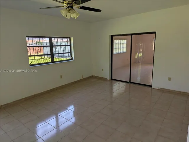 Neutral ceramic tile flooring, sliding glass doors, window with decorative security bars, and a ceiling fan with integrated light fixture