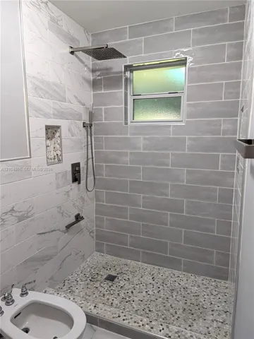 Walk-in shower featuring gray subway-style wall tile, contrasting white stone-finish wall accents, a pebble-finish floor, built-in recessed niche, and metallic rainfall showerhead with a separate hand-held sprayer