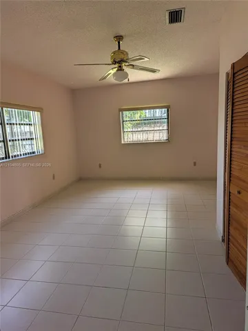 Interior space featuring neutral tile flooring, louvered wood-finish closet doors, multi-pane windows with metal security bars, and a ceiling-mounted light fixture with a fan