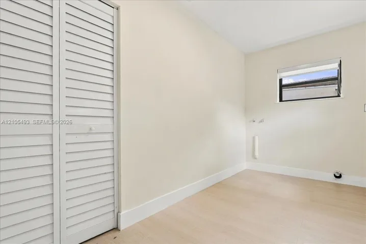 Neutral-toned interior room featuring wood-finish flooring, white louvered closet doors, a horizontal window with integrated roller shade, and wall-mounted utility connections