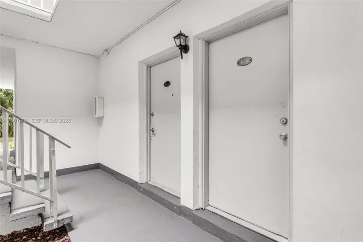 Covered entryway featuring white exterior walls, painted concrete flooring, a metal staircase with railing, and wall-mounted exterior lighting