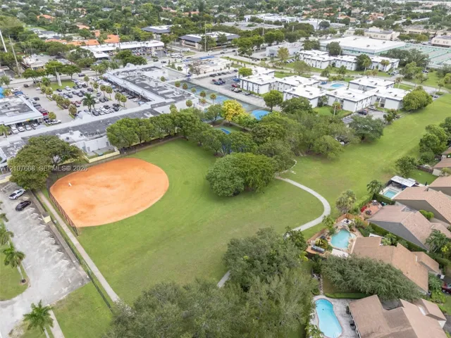 Aerial view of a sprawling recreational area featuring an expansive grass field, a dedicated baseball diamond, paved walkways, and mature trees