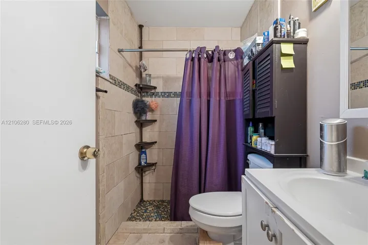 Bathroom featuring tile-surround shower stall with mosaic accent inlay, neutral large-format wall tile, and white vanity with integrated sink