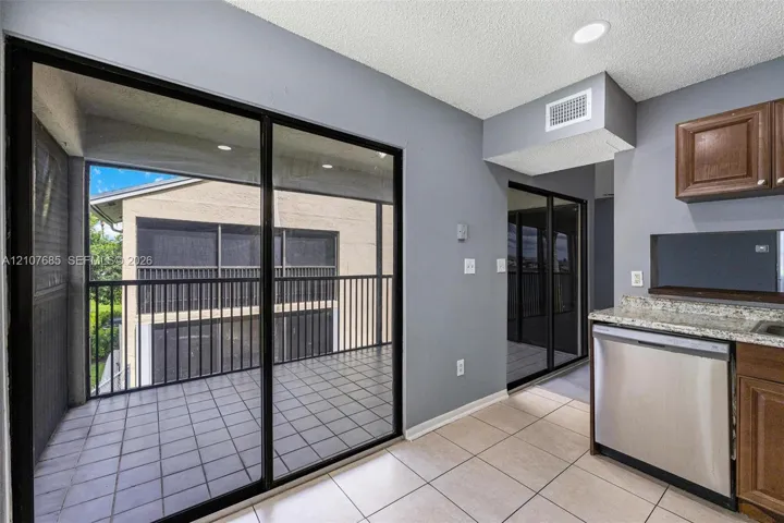 Sliding glass doors leading to a covered balcony with tiled flooring and metal railing, paired with an interior kitchen featuring stainless steel dishwasher, stone-finish countertops, and wood-finish cabinetry