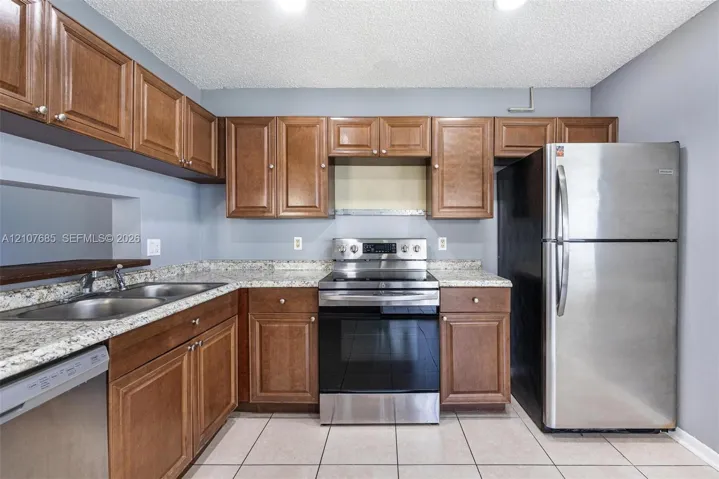 Kitchen featuring raised-panel wood cabinetry, stone-patterned countertops, stainless steel appliances, a double-basin stainless steel sink, and ceramic tile flooring