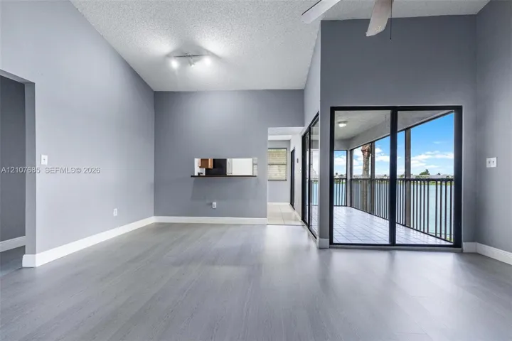 Vaulted ceilings featuring a ceiling fan, neutral wall paint, wood-finish flooring, a kitchen pass-through window, and floor-to-ceiling glass sliding doors opening to a covered balcony