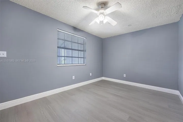 Room featuring wood-finish flooring, textured ceiling, light-toned wall paint, white-framed window with blinds, and a white ceiling fan with integrated light fixture
