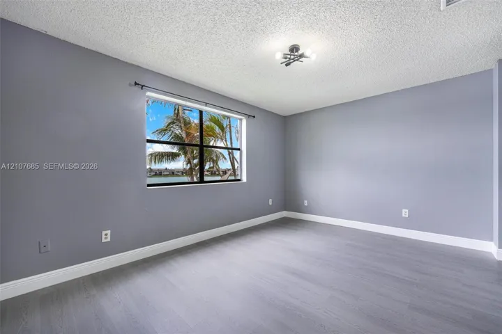 Room featuring gray-tone walls, wood-finish flooring, a multi-bulb flush-mount light fixture, and a single-pane window with exterior water and palm tree scenery
