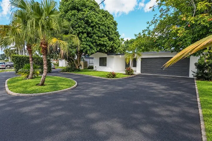 Single-story residence featuring an expansive asphalt driveway, a multi-car garage with paneled door, integrated landscaping with mature palm trees, and manicured lawn edging