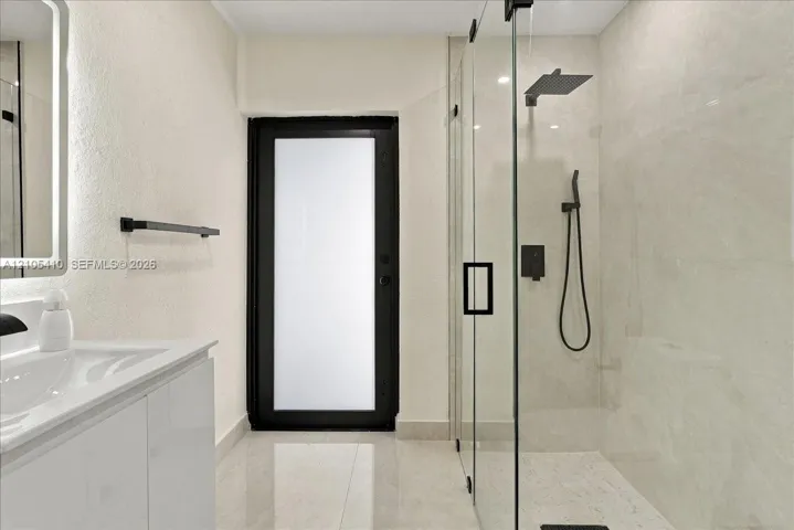 Bathroom featuring a walk-in shower with frameless glass enclosure, matte black rainfall showerhead, and handheld wand, accompanied by a white vanity with integrated sink and a frosted glass exterior door