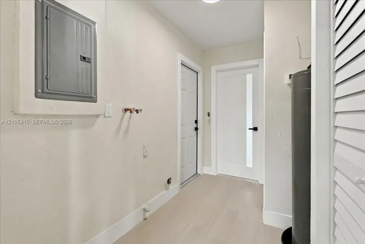 Utility area featuring wood-finish flooring, a metal electrical service panel, water hookups, and a white interior door with a vertical glass panel