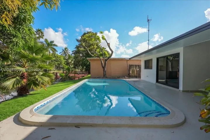 In-ground swimming pool with a concrete surround, covered patio, manicured lawn, and mature palm trees