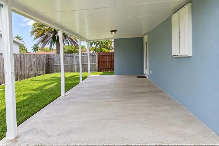 Covered concrete patio featuring a solid roof, white support pillars, exterior wall-mounted shutter, and a fenced backyard with lawn