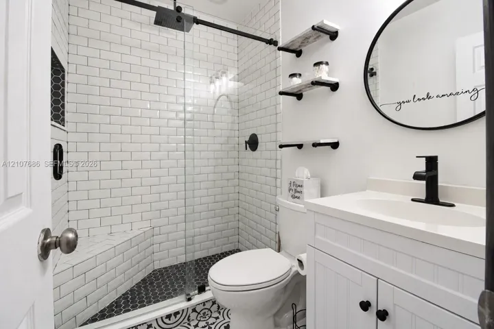 Bathroom featuring a glass-enclosed shower with white subway tile, hexagonal floor tiling, a white vanity with a single sink, matte black hardware, and wall-mounted shelving