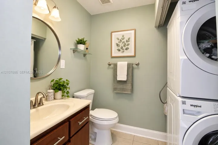 Laundry area featuring a stackable washer and dryer, wood-finish vanity with an integrated basin, circular wall mirror, and glass shelving