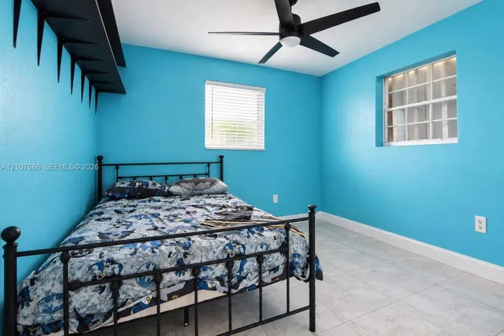 Neutral-toned tile flooring, matte black ceiling fan, dual windows with horizontal blinds, and a built-in wall-mounted shelf
