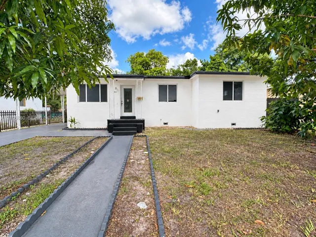 Single-story white brick exterior with a dark-finished entry staircase, central glass-paneled door, concrete walkway, and metal perimeter fencing