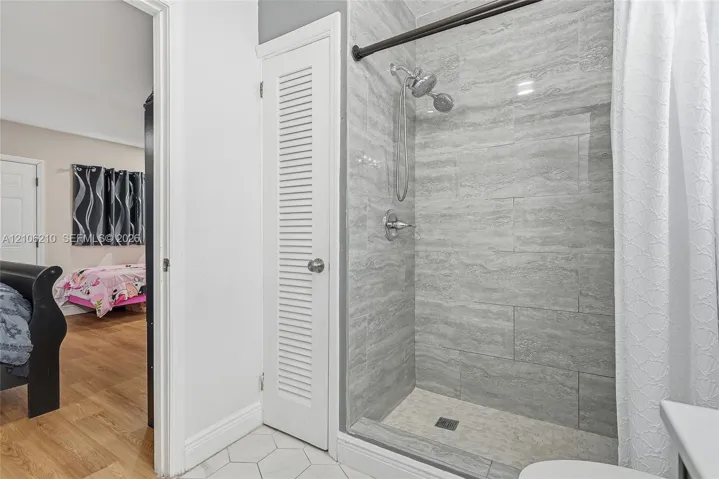 Walk-in shower with grey horizontal tile surround, handheld shower head, and built-in closet with white louvered door