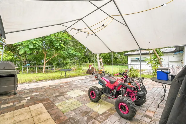 Paver patio area featuring a metal-framed canopy structure, chain-link perimeter fencing, and mature trees