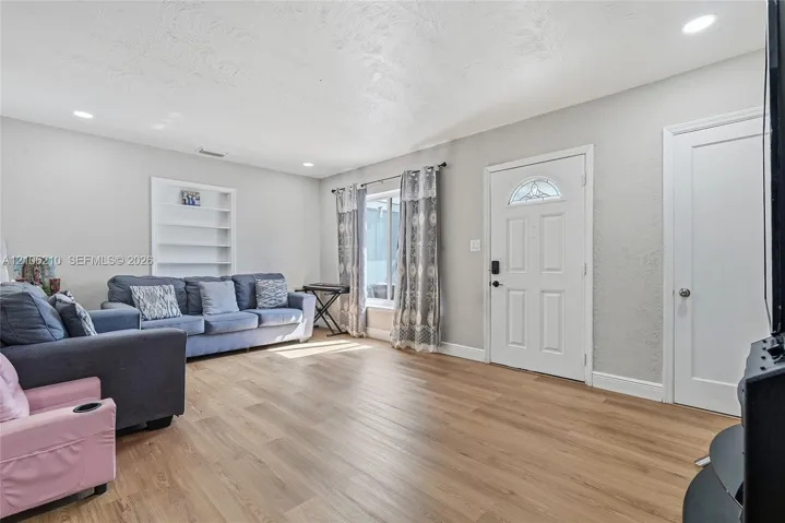 Living area featuring wood-finish flooring, recessed lighting, built-in wall shelving, and a paneled entry door with decorative glass insert