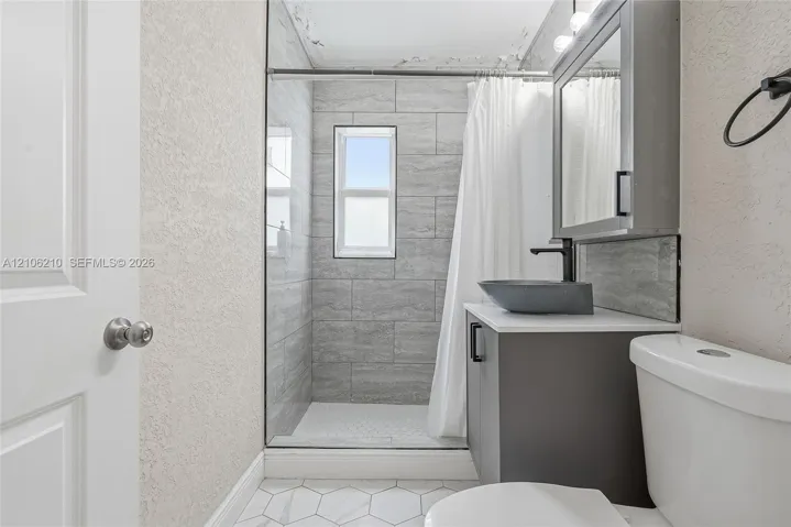 Bathroom featuring a shower stall with gray horizontal tile surround, a vanity with a dark cabinet and white countertop, a vessel sink with a matte black faucet, and white hexagonal floor tiling