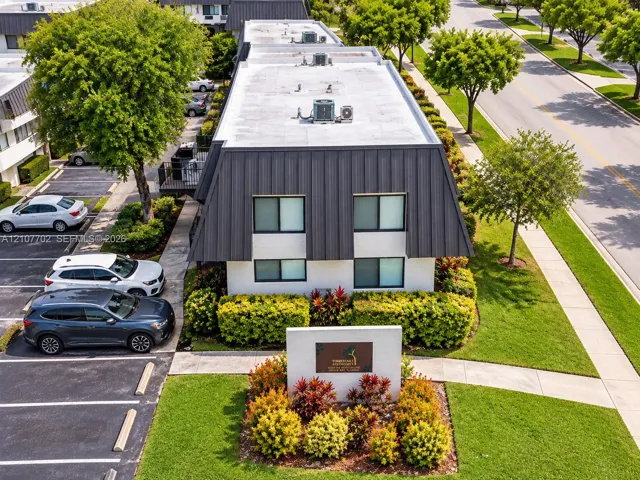 Exterior view of a multi-level building with metal mansard roof detailing, white stucco siding, dedicated surface parking, and curated landscaping